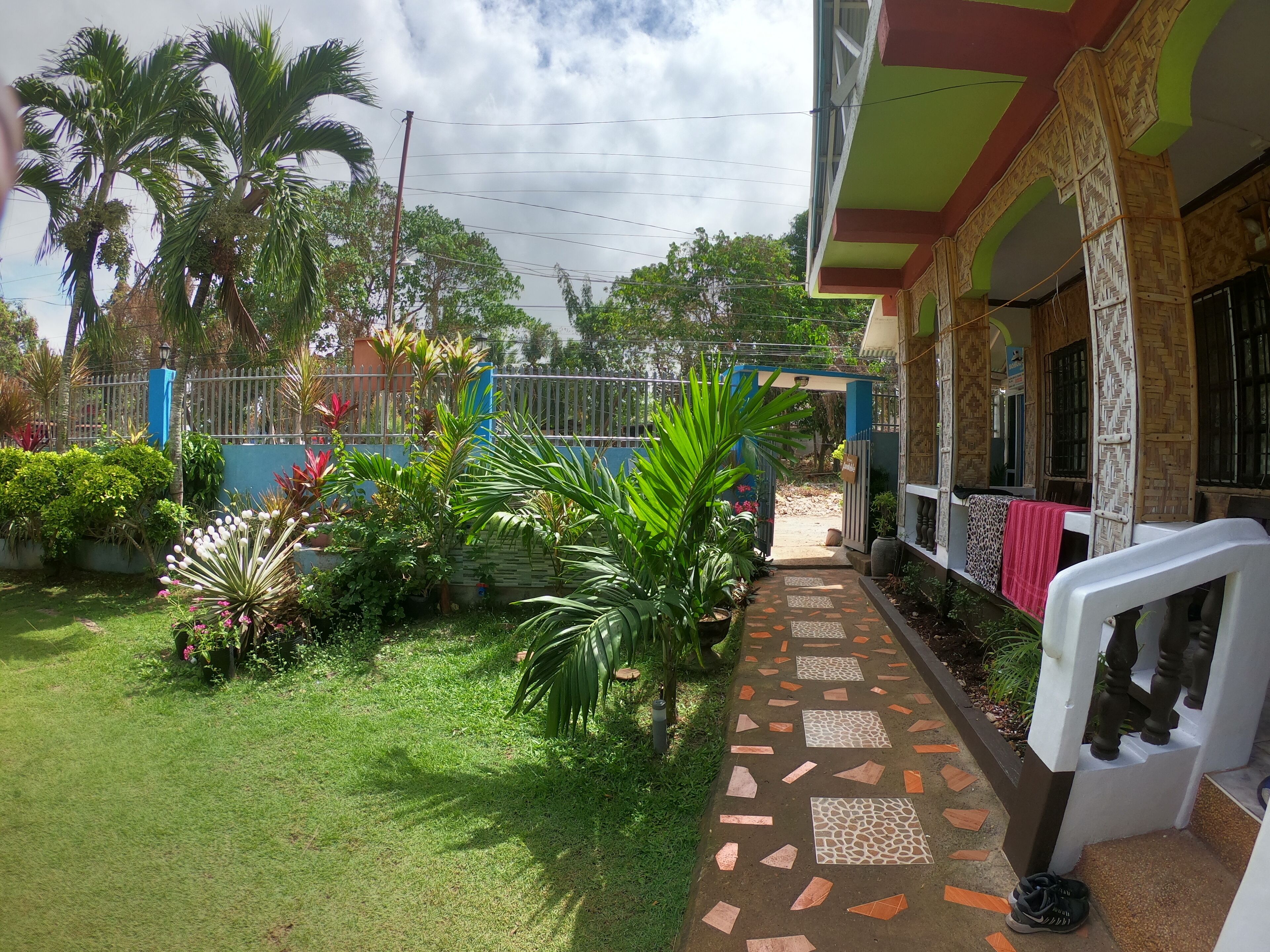Un jardín bien cuidado, con palmeras y diversas plantas, un camino empedrado que lleva a un edificio de diseño tradicional, y un área cercada.