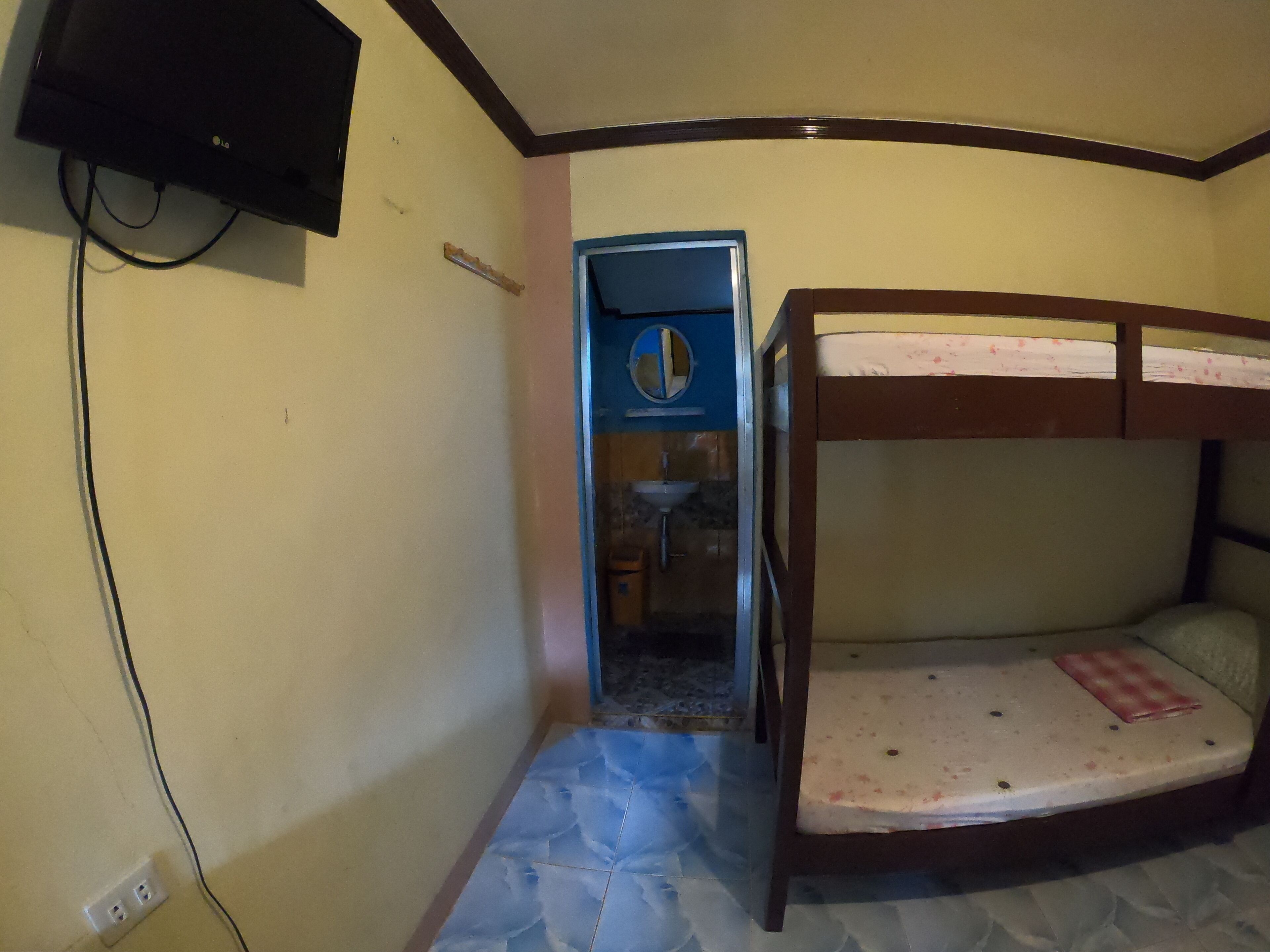A small room with a bunk bed, a wall-mounted TV, a bathroom visible through a door, and a tiled floor.