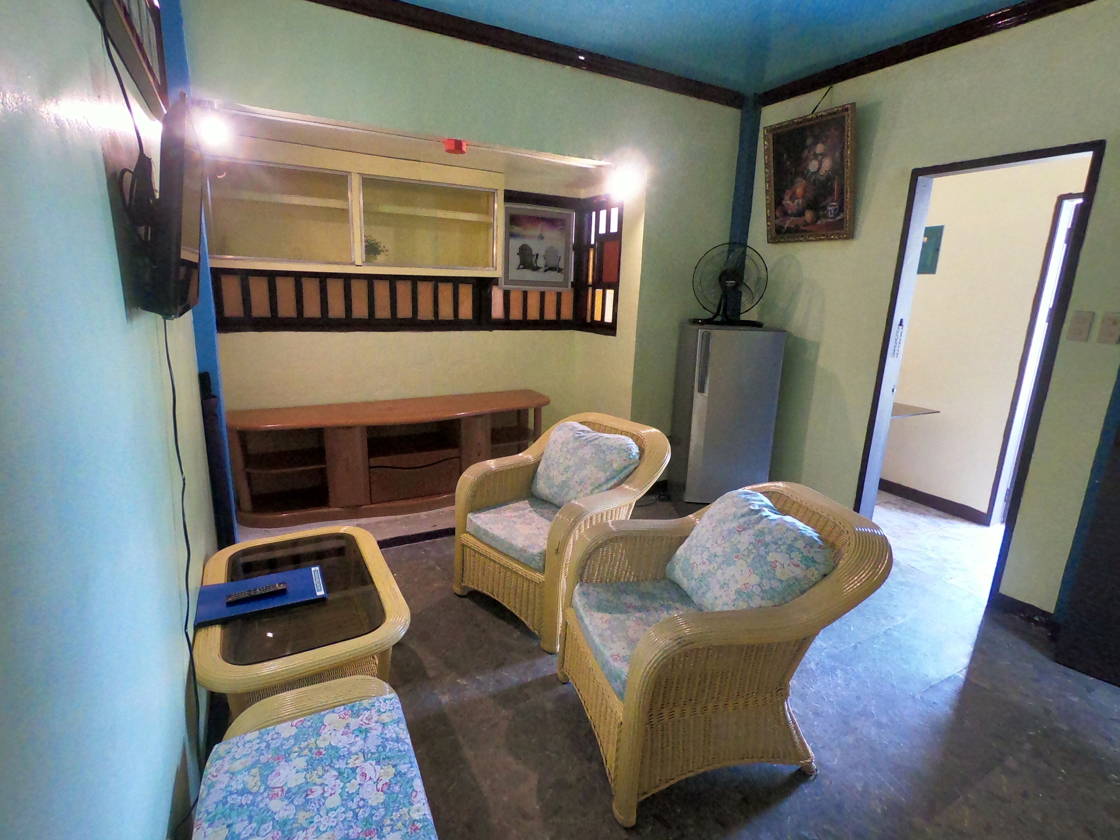 Una habitación con muebles de mimbre, un banco, una mesita y un refrigerador.