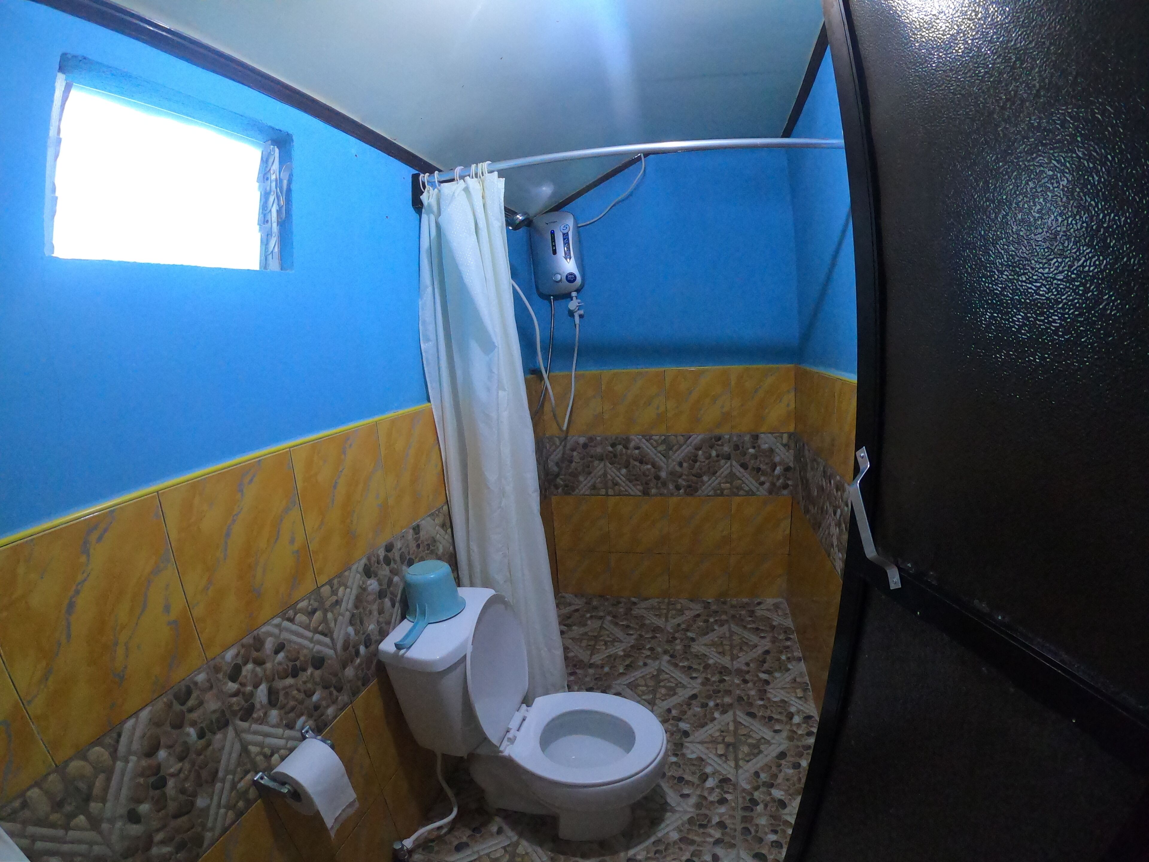 A bathroom with a blue and yellow tiled wall, a white toilet, a shower curtain, and a small window.