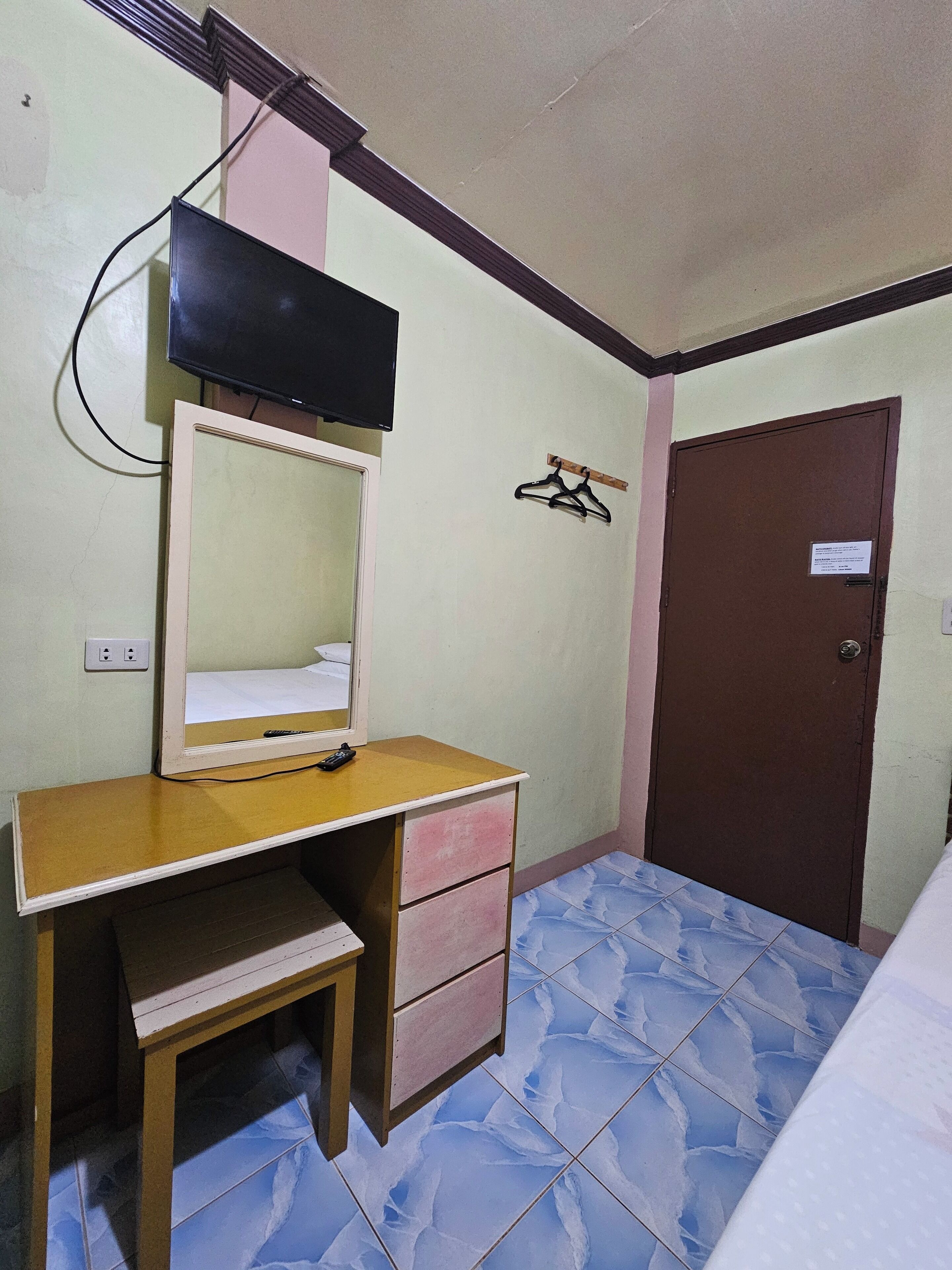 A small bedroom with a wooden desk, a mirror, a television mounted on the wall, a door, and a tiled floor.