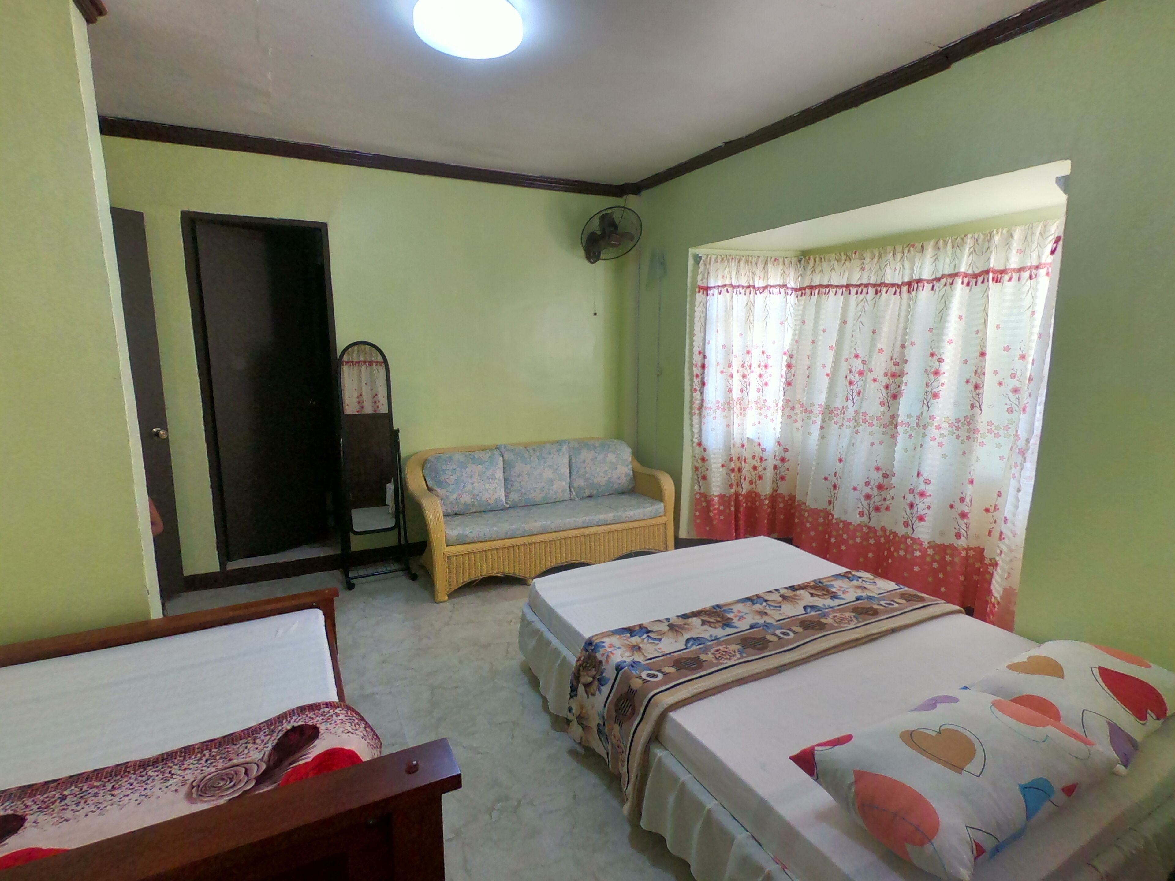 A room with two single beds, a sofa, a chair, a fan, and a window with floral curtains.