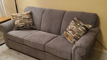 A grey sofa with two patterned cushions, a wooden floor, and a wall with a horizontal wooden panel and a painting.