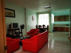 A living room with a red sofa, a TV, and a fan. There is a dining area with chairs and a table. - Villa Lolita Apartelle (Tacloban)