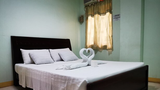 Un letto rifatto con lenzuola e cuscini bianchi, un asciugamano disposto a forma di cuore e una testiera in legno scuro.