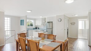 A modern kitchen with white cabinets, stainless steel appliances, and a dining area with wooden chairs and a table. - Luxury Boardwalk Apartment - Unit 7 (NAROOMA)