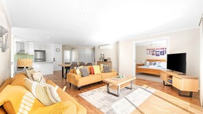 A modern living room with a sofa, coffee table, and a TV. There is a dining area with chairs and a table. The kitchen is visible in the background with cabinets and appliances. - Luxury Boardwalk Apartment - Unit 7 (NAROOMA)