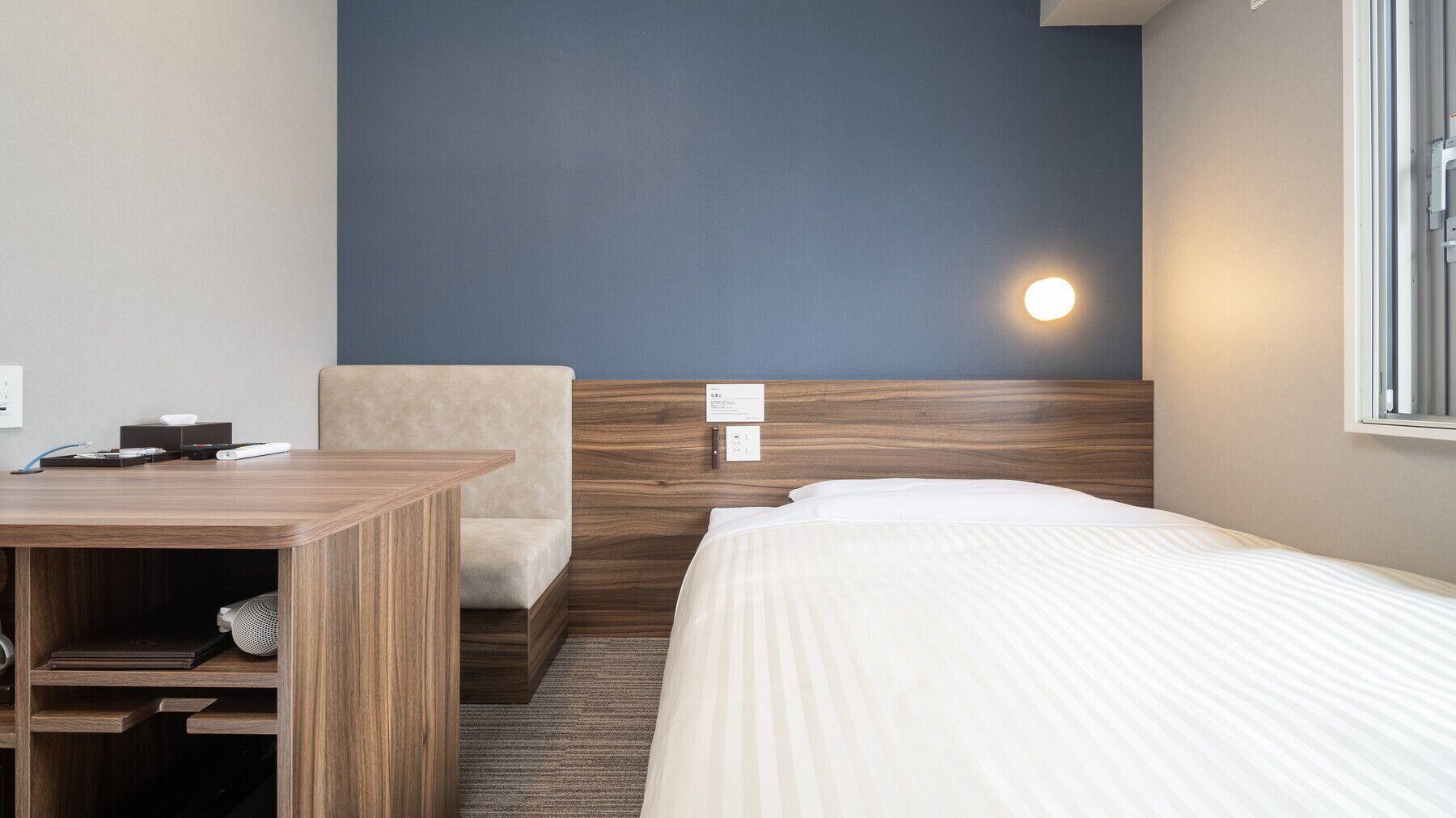 A hotel room with a wooden desk, a chair, a bed, a nightstand, and a wall-mounted light.