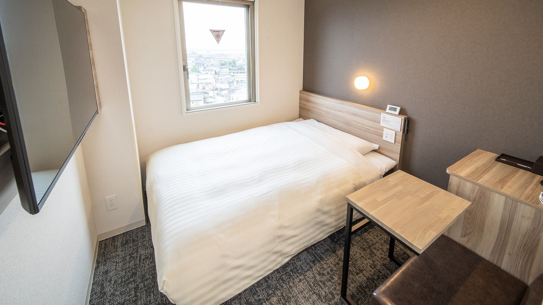 A hotel room with a bed, a small table, a nightstand, a window with a view, and a wall-mounted light.