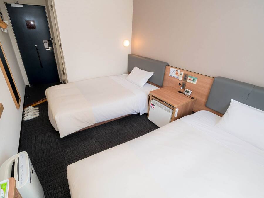 A hotel room with two single beds, a small bedside table with a lamp, a mini fridge, and a door with a handle.