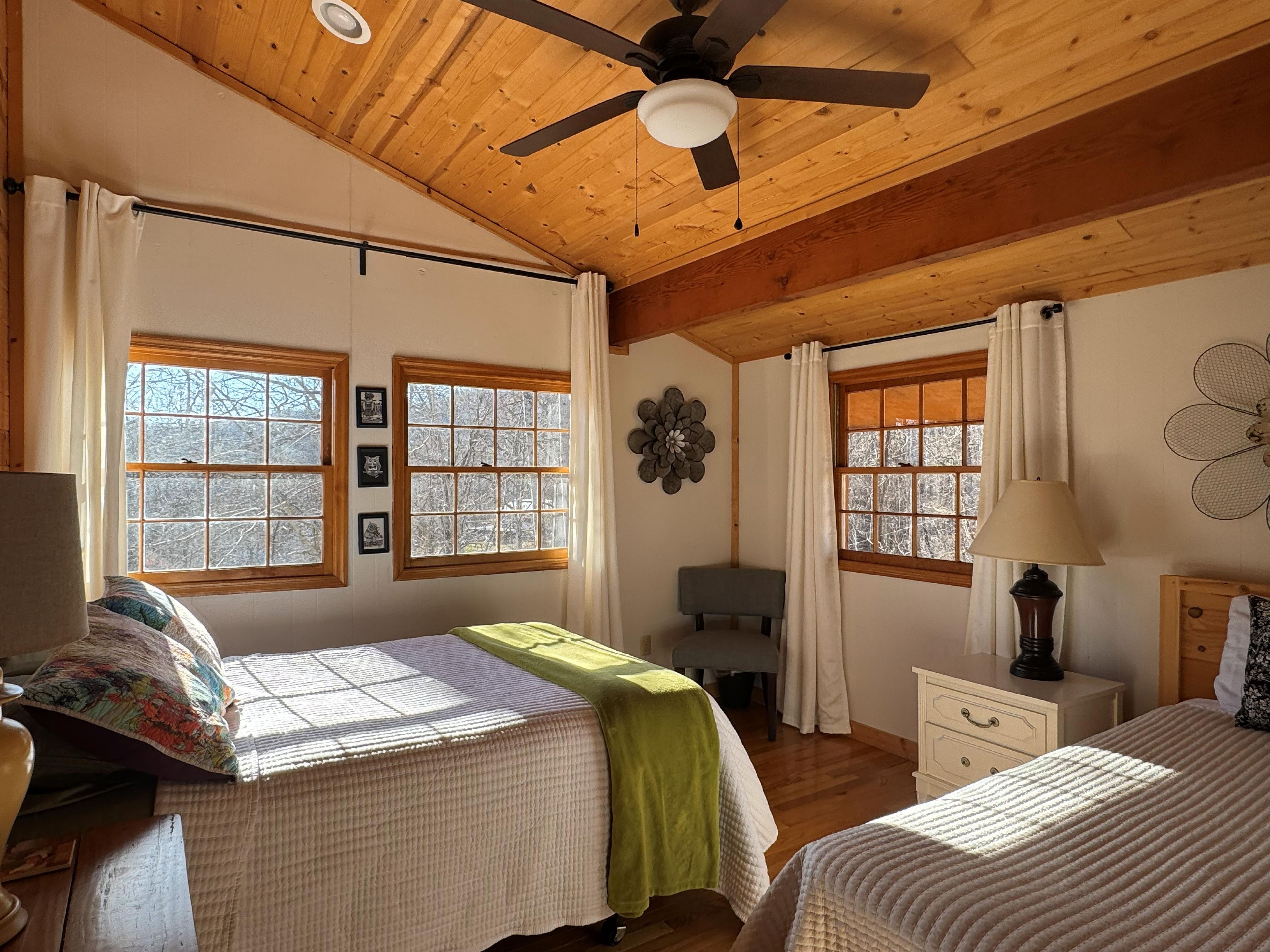 A bedroom with two beds, wooden ceiling beams, a ceiling fan, and large windows with curtains.