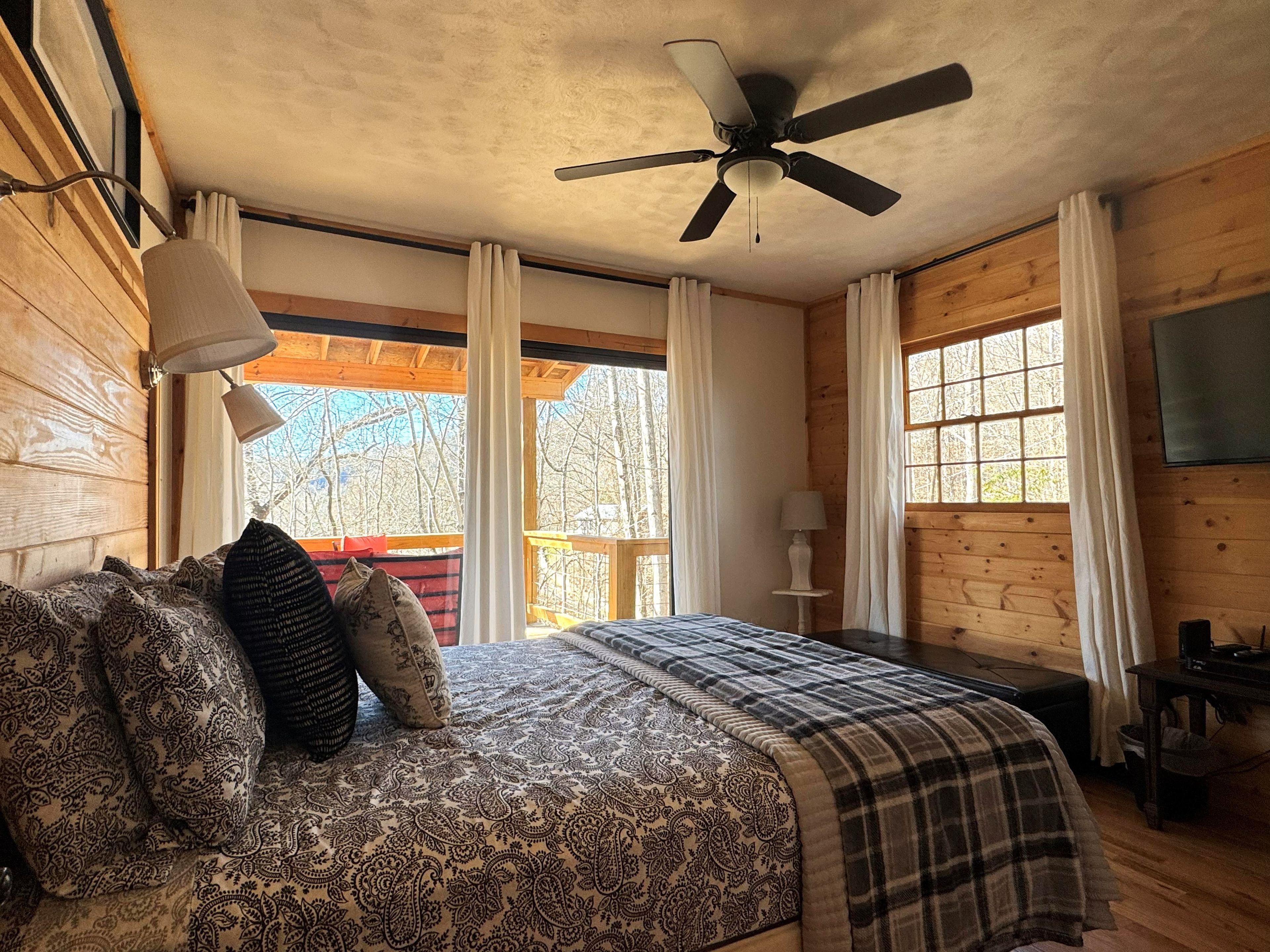 Une chambre à coucher avec un lit, un ventilateur de plafond, une fenêtre avec des rideaux et un téléviseur fixé au mur.