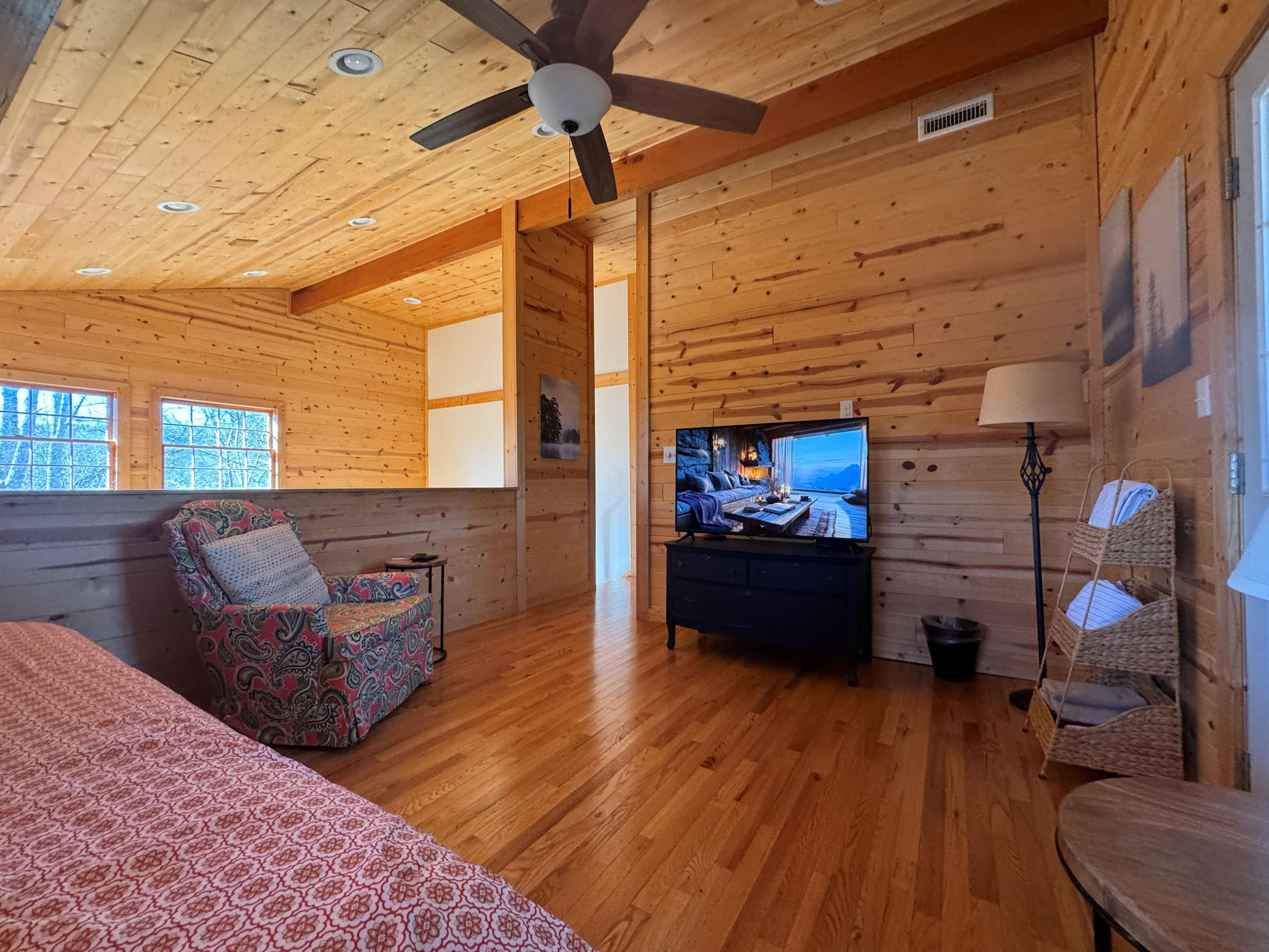 A wooden cabin interior with a bed, a patterned armchair, a television, and a ceiling fan.