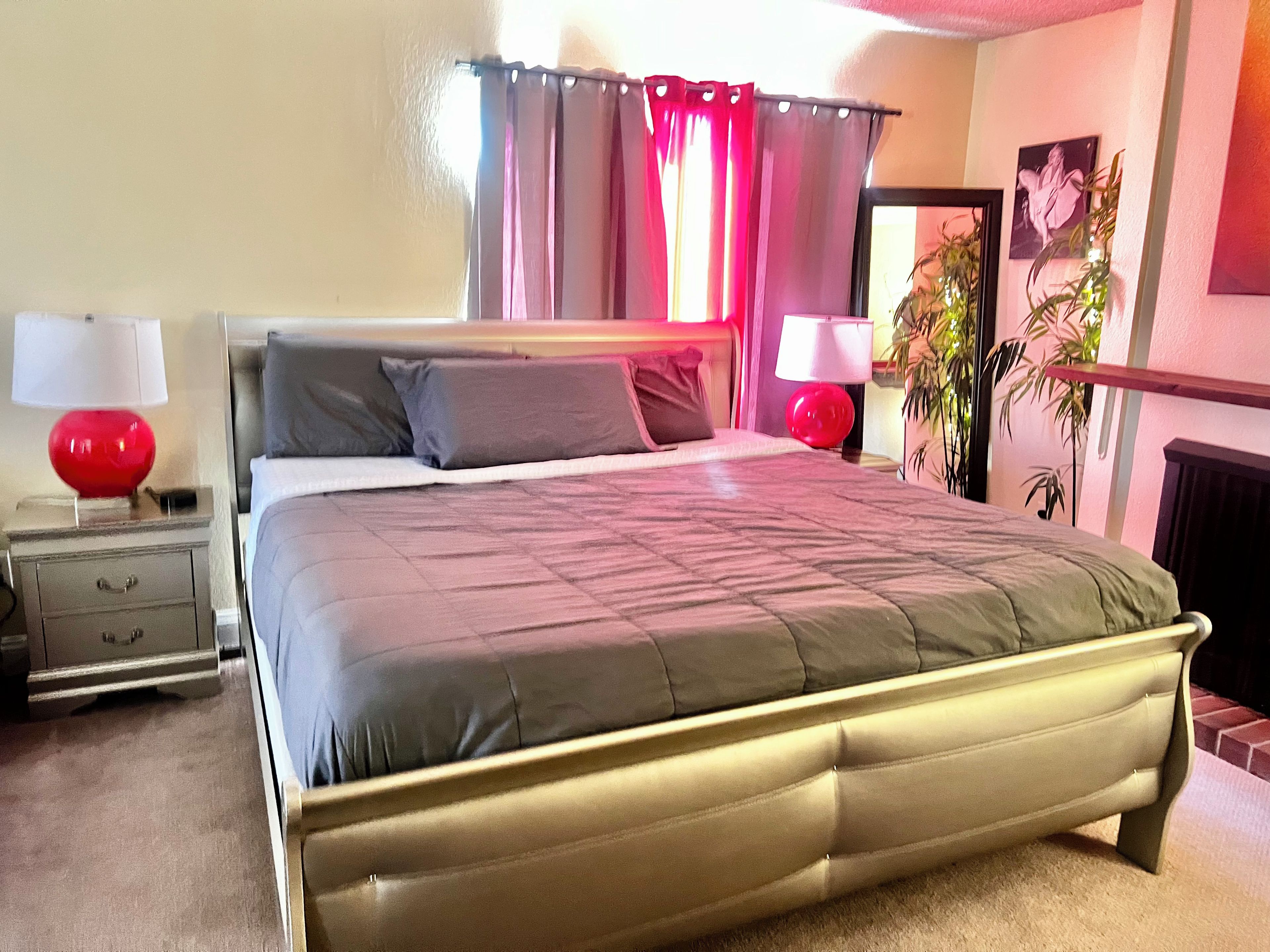 Een slaapkamer met een bed, twee nachtkastjes, een lamp, een ronde rode lamp, een rood gordijn, een aan de muur hangend schilderij en een plant in een pot.