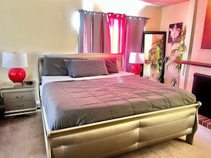 Un dormitorio con cama, dos mesitas de noche, una lámpara, una lámpara esférica roja, una cortina roja, un cuadro colgado en la pared y una planta en maceta.