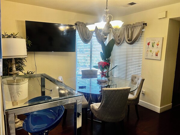 A dining area with a glass table, chairs, a blue bowl, a TV mounted on the wall, and a chandelier. - Prime Location! Cozy home with pool, just minutes from the airport & Strip! (Las Vegas)