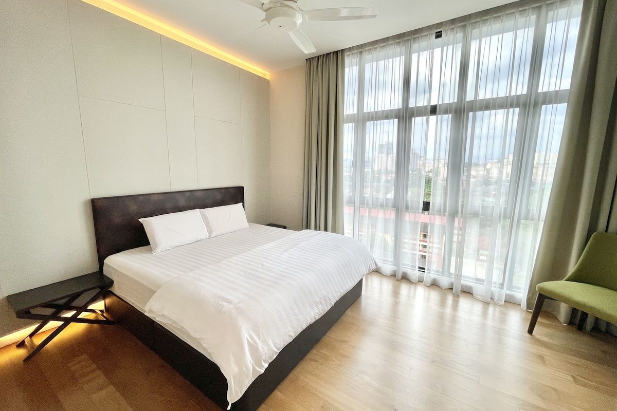 Une chambre d’hôtel moderne avec un grand lit, un ventilateur de plafond et une vue sur la ville à travers de fines rideaux.