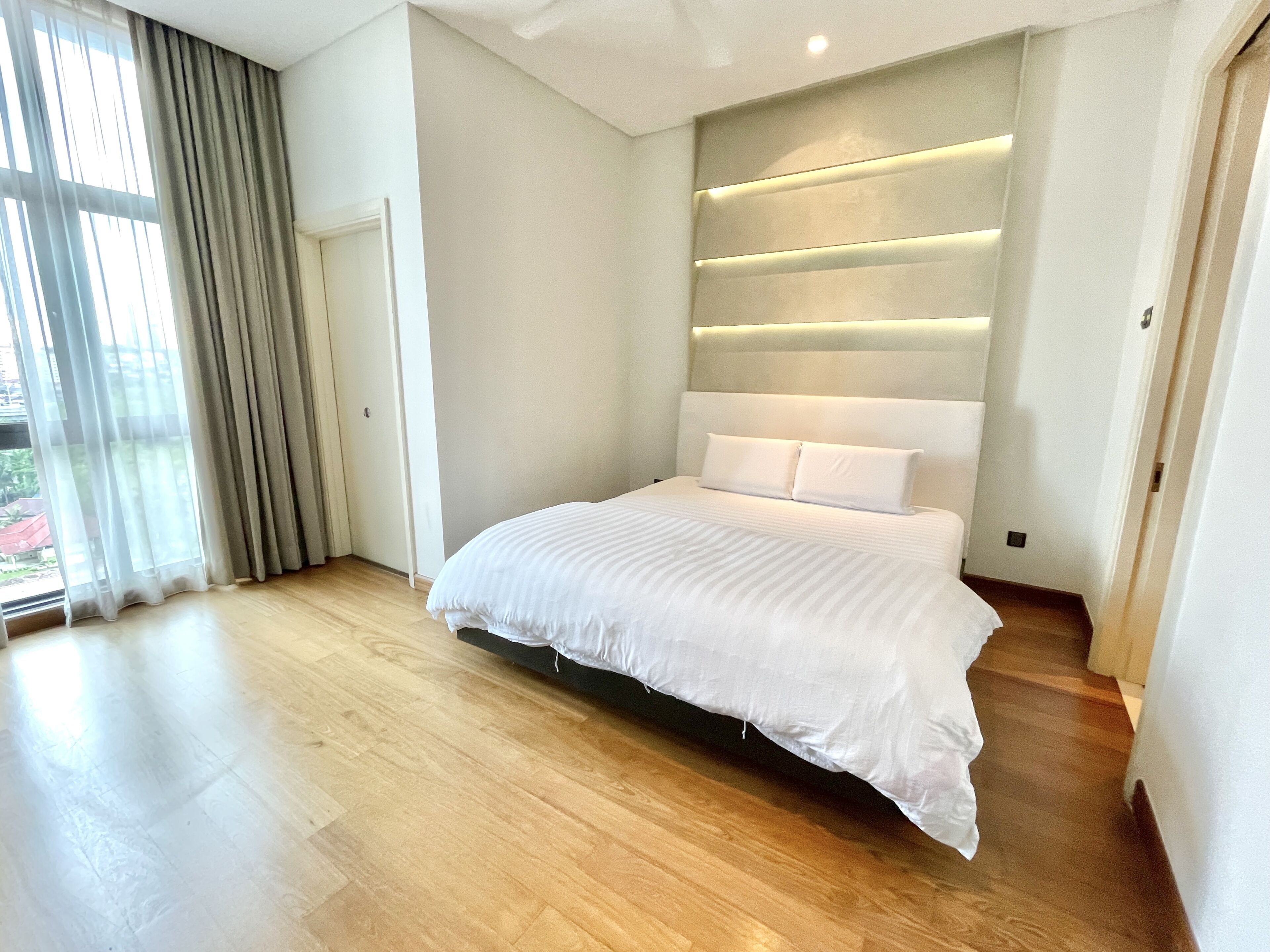 Un dormitorio moderno con una cama grande, suelo de madera y vistas al exterior a través de una ventana con cortinas.