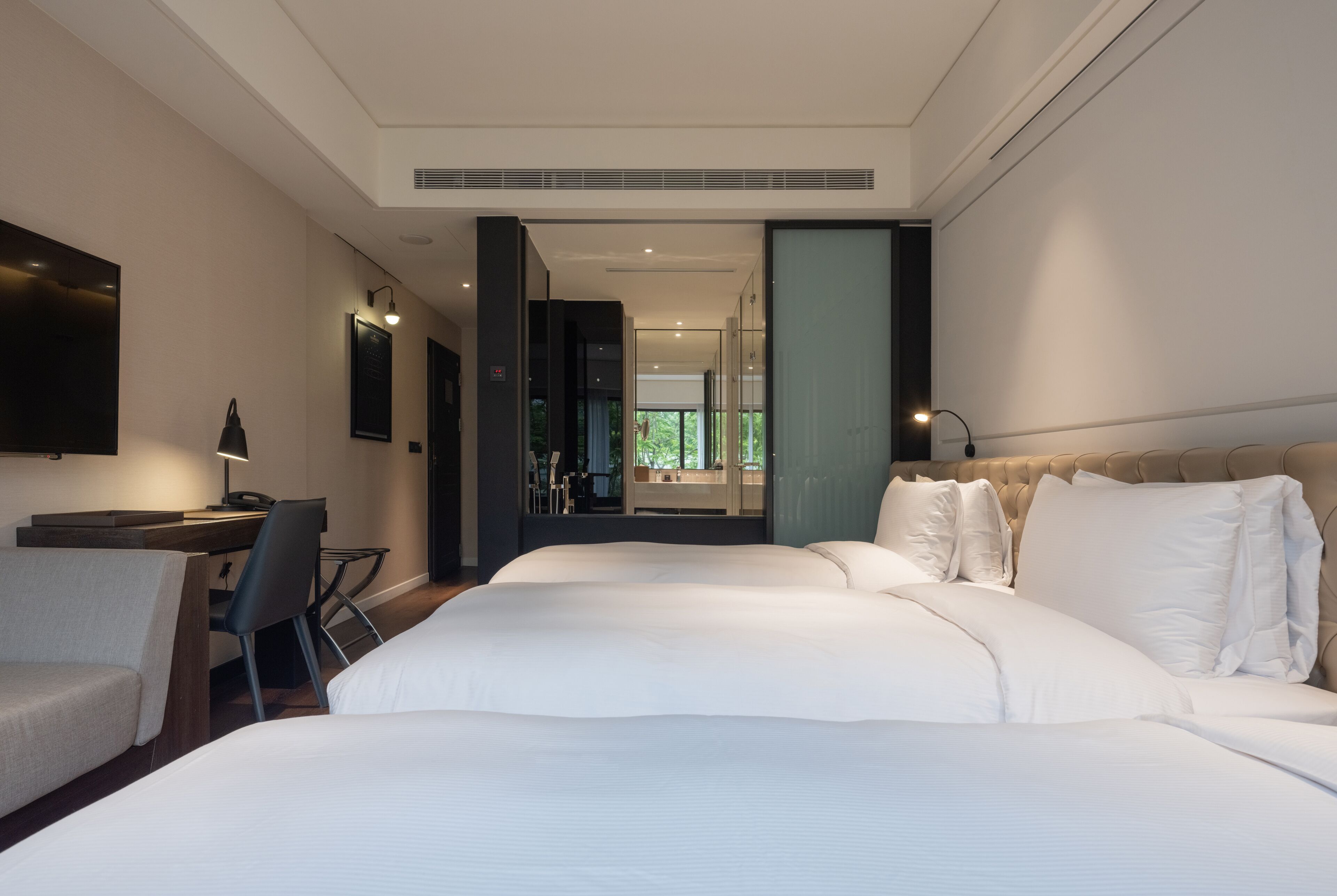 A modern hotel room with a large bed, a desk, and a chair. There is a TV mounted on the wall and a window providing a view of the outdoors.