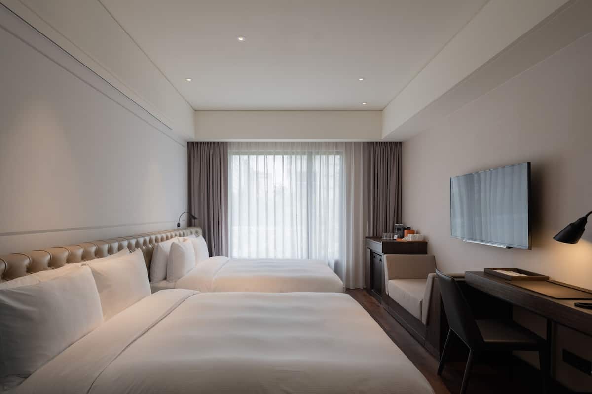 Una habitación de hotel con dos camas, un escritorio, una silla, un televisor y una ventana con cortinas.