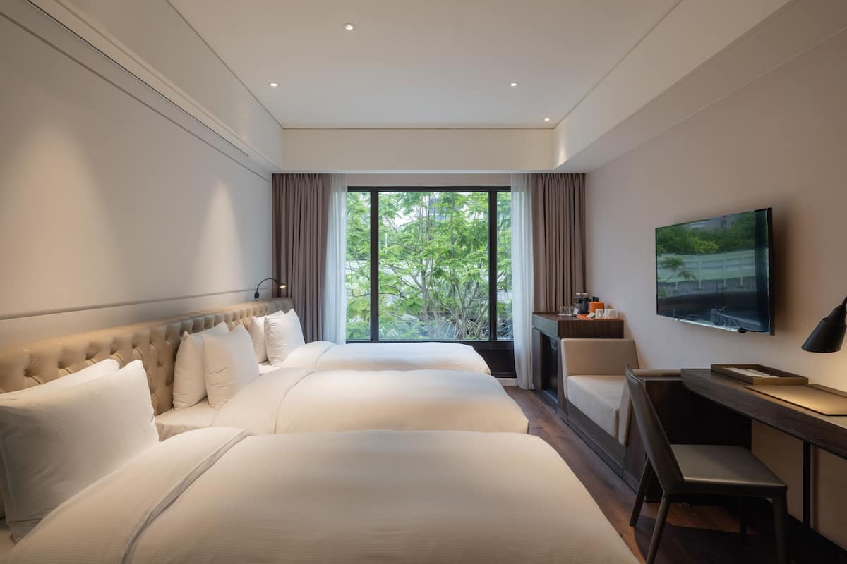 Habitación de hotel con dos camas, un escritorio con silla, televisor y ventana con cortinas.