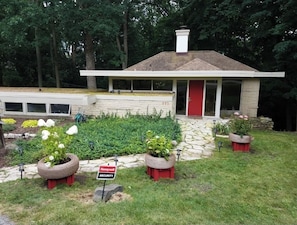 Uma casa moderna com uma porta vermelha, rodeada por um jardim com vasos de plantas e uma placa de segurança.