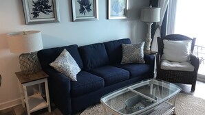 Een woonkamer met een donkerblauwe bank, twee bijpassende kussens, een glazen salontafel, een bijzettafeltje met een lamp, en drie ingelijste kunstwerken aan de muur.