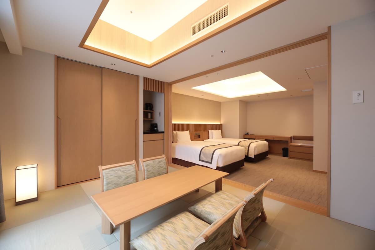 A modern hotel room with a wooden dining table, two beds, a small kitchenette, and a sliding door closet.