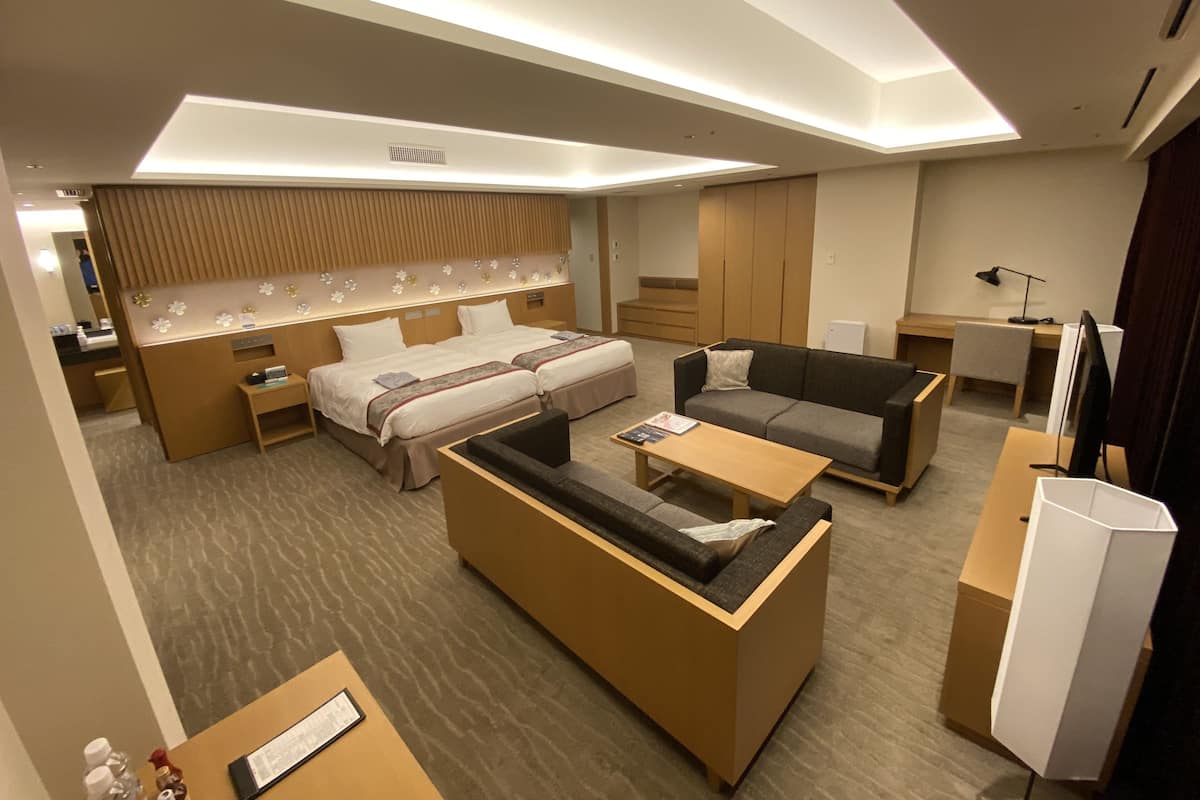 A modern hotel room with a sofa, two beds, a desk, and a flat-screen TV.