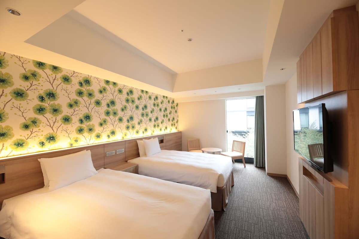 A hotel room with two beds, a TV, a small table, and a window with a view.