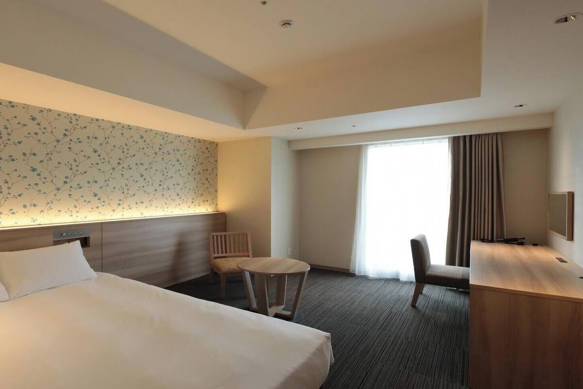 A hotel room with a large bed, a desk, a chair, a small table, a TV, and a balcony with curtains.
