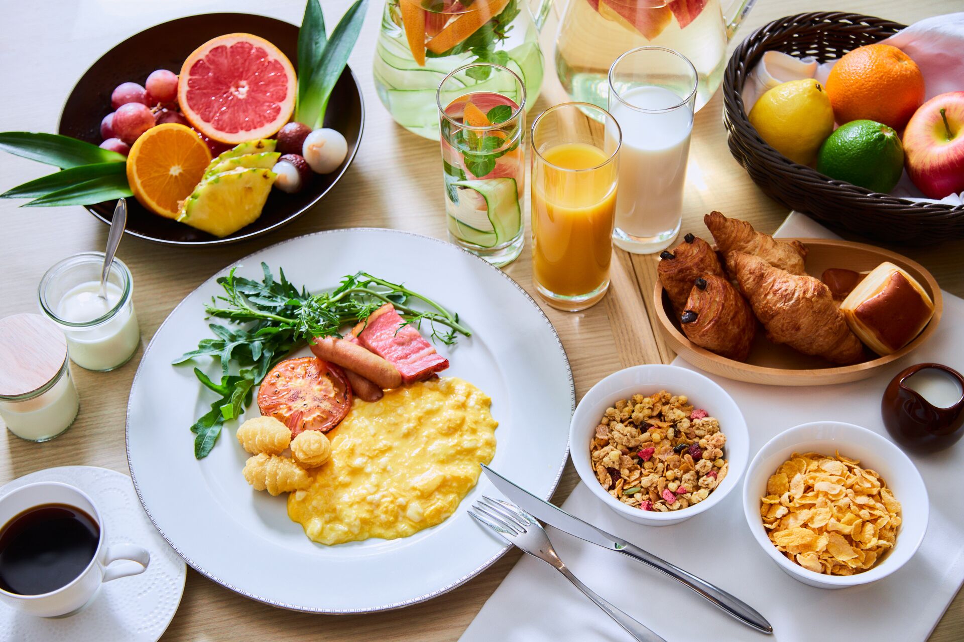 A breakfast setting with scrambled eggs, ham, tomatoes, and greens on a plate, accompanied by croissants, cereal, and fruit.