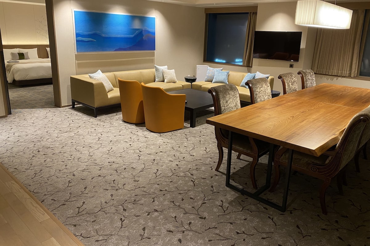 A hotel room with a large wooden dining table, a sofa set, a bed, and a large wall mural.