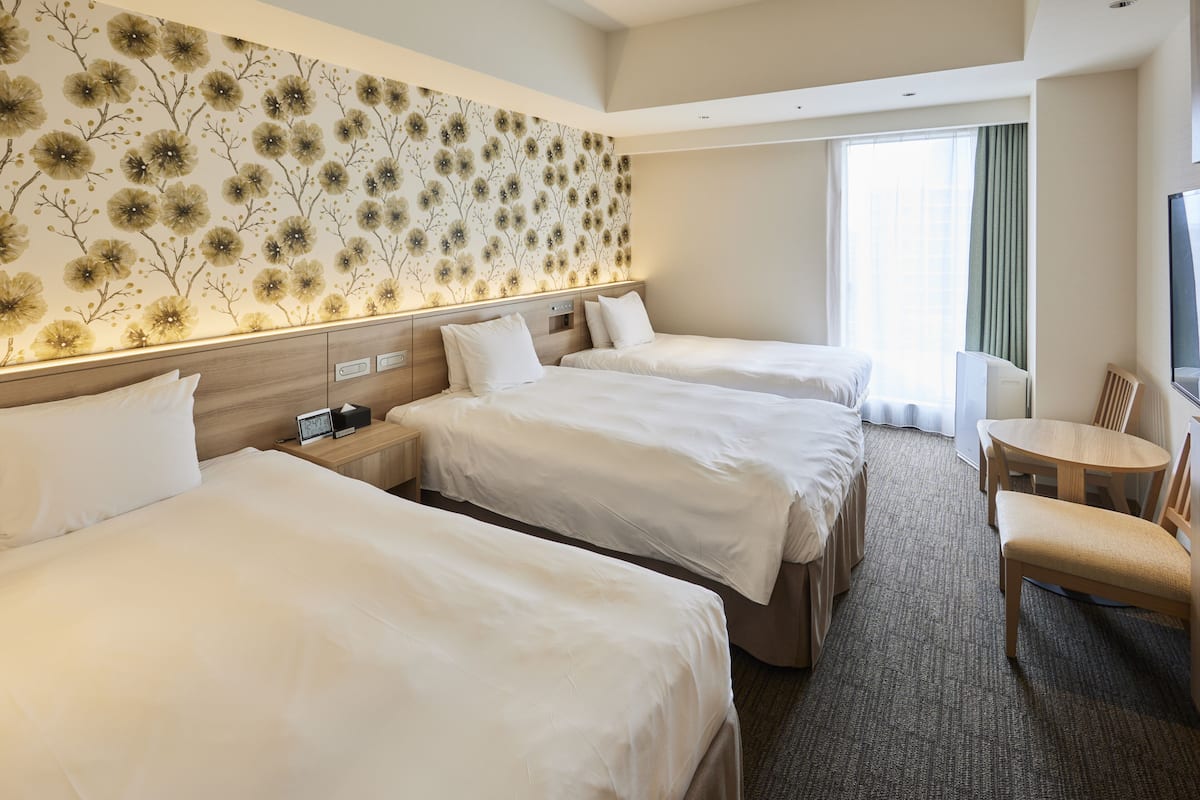 A hotel room with three beds, a wooden headboard, a wall-mounted TV, a desk with a chair, and a large window with curtains.