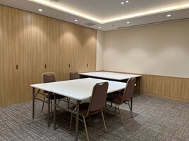 A conference room with two tables and four chairs, wooden panel walls, carpeted floor, and recessed ceiling lighting.