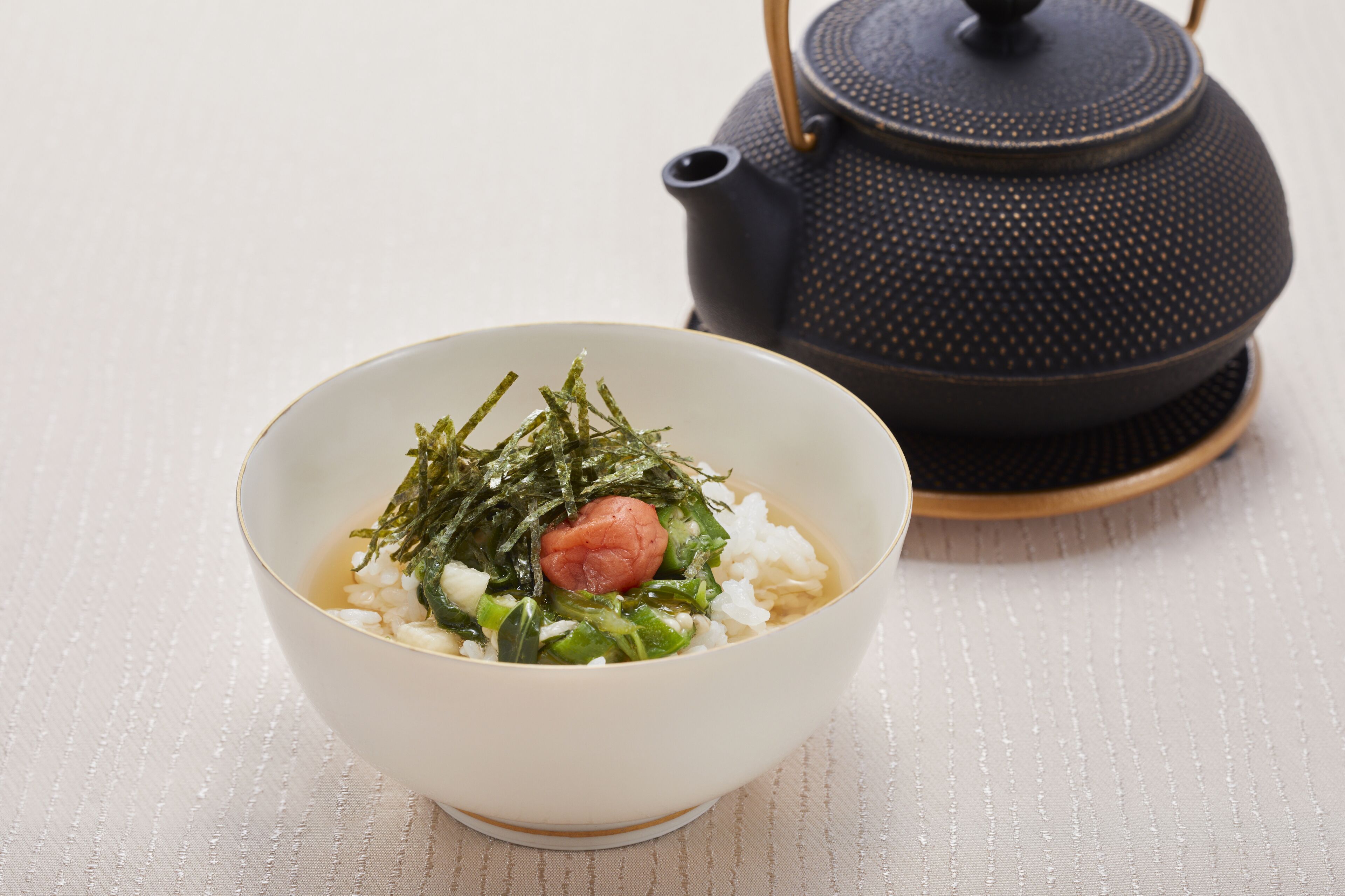 A bowl of rice with seaweed and a sausage, and a teapot in the background.