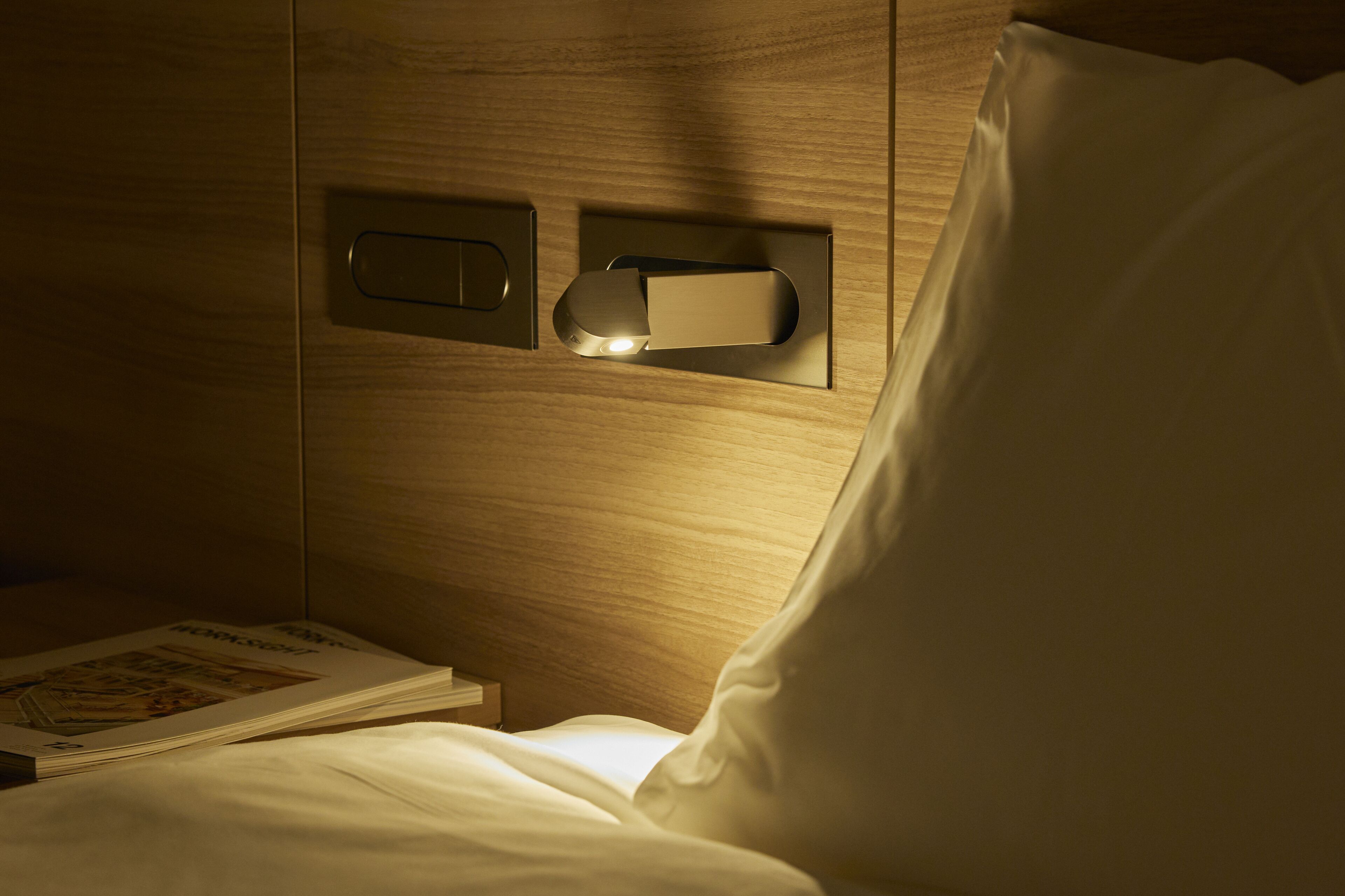 Quarto de hotel com cabeceira de madeira, mesa de cabeceira com uma revista e um criado-mudo.
