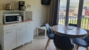 A small kitchen with a microwave, a coffee maker, and a TV on the wall. There is a dining table with chairs and a view of buildings through the window. - GRANDALLA  31 -1 apt 3 pers-Pas de la Casa- pie piste-006160 (PAS DE LA CASA)