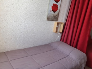 Ein Einzelbett mit grauer Bettdecke, ein Holzregal mit Steckdose und ein Bild von roten Blumen an der Wand.