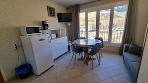 A compact kitchen with a refrigerator, microwave, and coffee maker. There is a dining table with chairs and a sofa. The room has a large window with a view of a mountainous landscape. - GRANDALLA  31 -1 apt 3 pers-Pas de la Casa- pie piste-006160 (PAS DE LA CASA)
