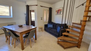 A room with a wooden dining table, chairs, a sofa with cushions, a wooden staircase, and a window with curtains.