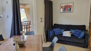 A living room with a dark grey sofa, blue cushions, a wooden dining table with a white vase and a basket, and a painting of flowers on the wall.