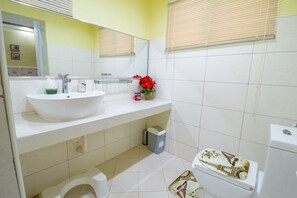 Ein Badezimmer mit weißem Waschbecken, Spiegel und Toilette. Ein Mülleimer und ein kleines Fenster mit Jalousien sind ebenfalls vorhanden.