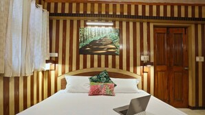 Une chambre d’hôtel avec un lit, un ordinateur portable posé sur le lit, un tableau représentant un sentier forestier accroché au mur et une porte en bois.