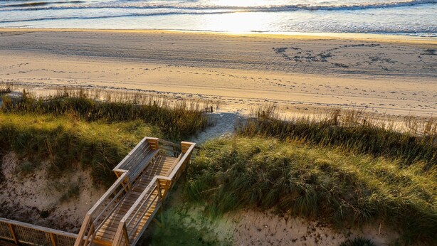 A beach with a wooden boardwalk leading to the water, surrounded by grass and sand.