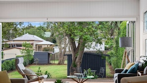 A spacious living room with a glass coffee table, comfortable seating, and a view of a backyard with a tree and a fence. - The Keeper's House (East Toowoomba)