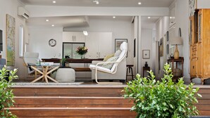 A modern living room with a wooden staircase, a white armchair, a glass coffee table, and a dining area with a kitchen in the background. - The Keeper's House (East Toowoomba)