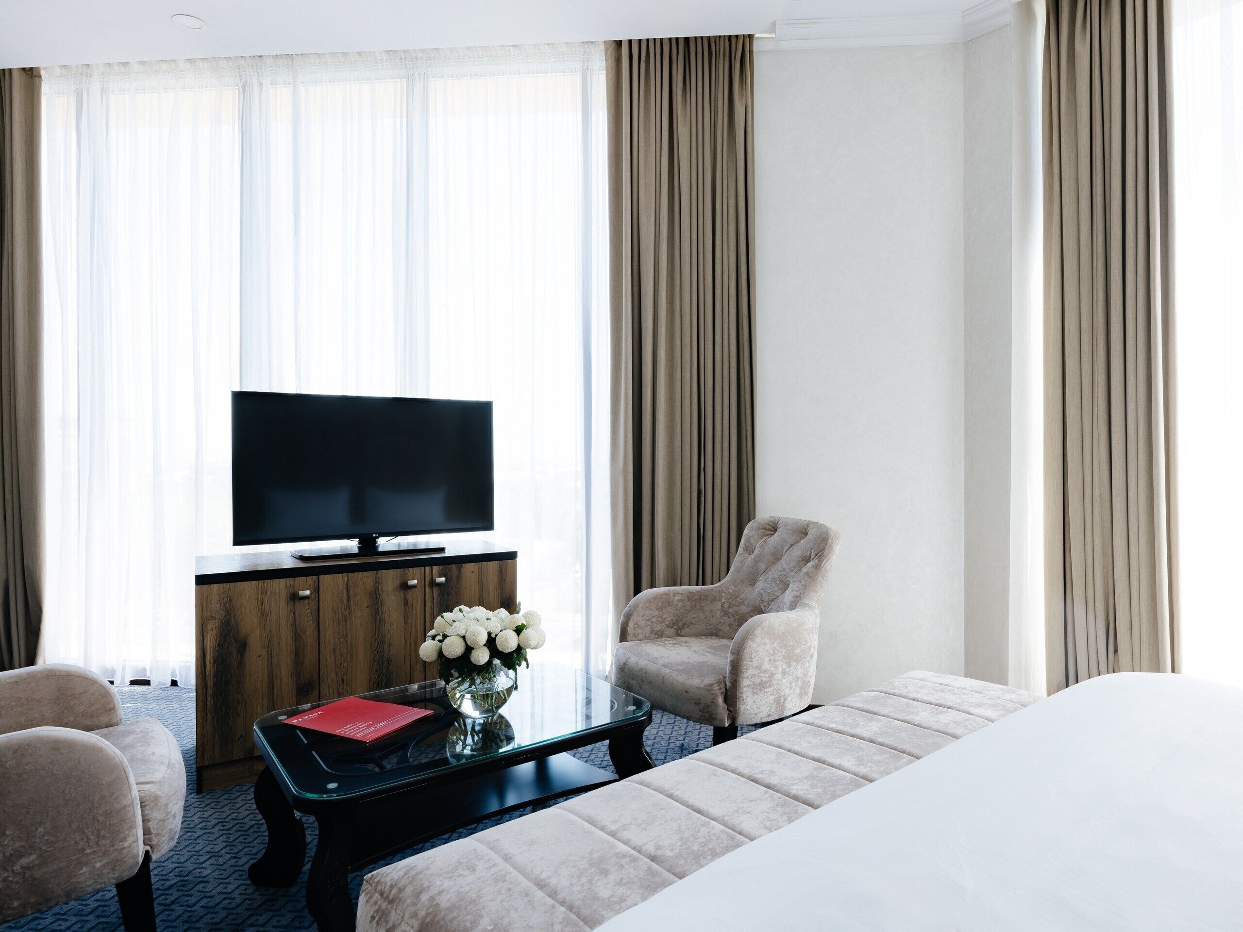A hotel room with a bed, a TV on a wooden cabinet, a chair, a coffee table with a vase of flowers, and curtains.