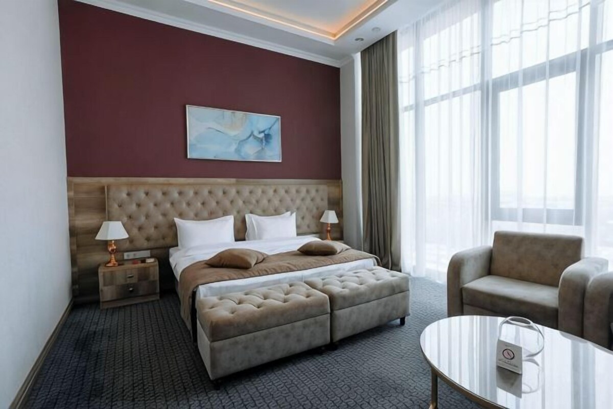 Habitación de hotel con una cama grande, dos mesitas de noche, un sofá, una pequeña mesa y un ventanal amplio.