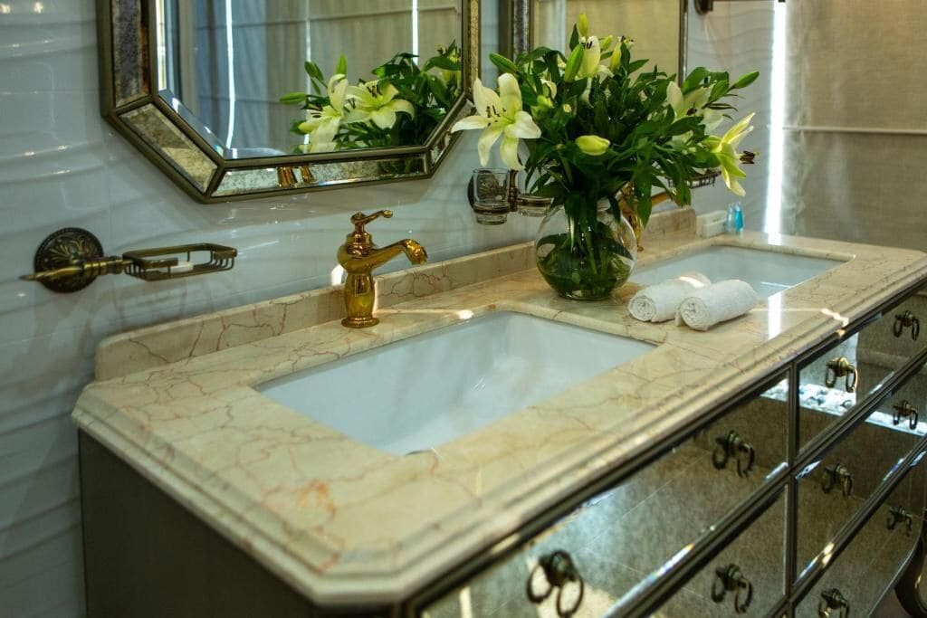 Un baño con lavamanos de mármol, grifería dorada, un espejo y un florero con flores.